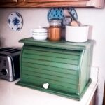 A rustic green bread box sits on a kitchen counter, topped with a ceramic jar, spice jar, and kitchen utensils. A toaster and a decorative plate are nearby.