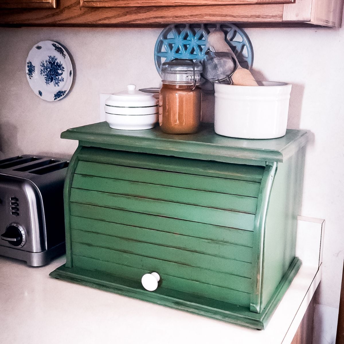 A rustic green bread box sits on a kitchen counter, topped with a ceramic jar, spice jar, and kitchen utensils. A toaster and a decorative plate are nearby.