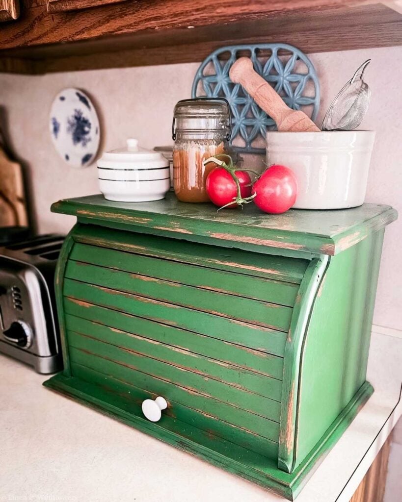 Rustic kitchen counter with a distressed green bread box, topped with tomatoes, honey jar, and utensils. Cozy and vintage vibe.