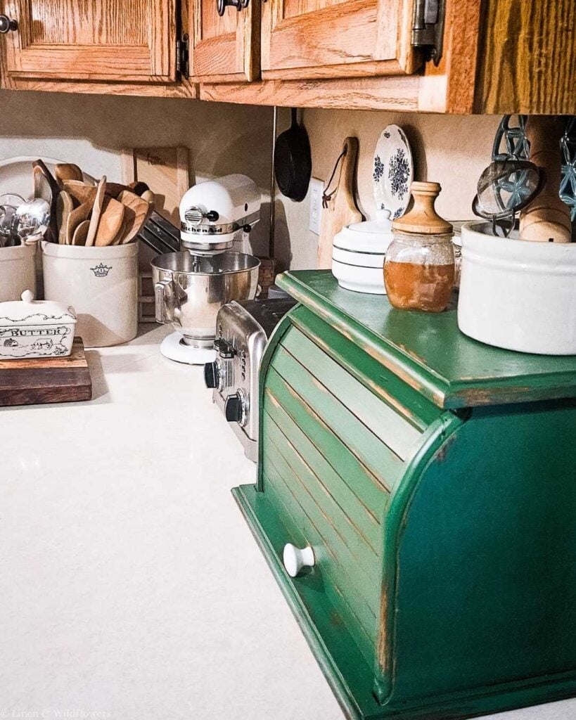 Cozy kitchen scene with rustic wooden cabinets, a vintage green bread box, cream-colored pottery, wooden utensils, and a white mixer on a countertop.