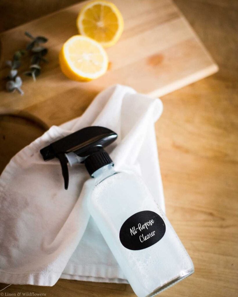 Homemade all-purpose cleaner spray bottle with black label on a white cloth next to sliced lemons and eucalyptus — frugal living cleaning tips for homemakers