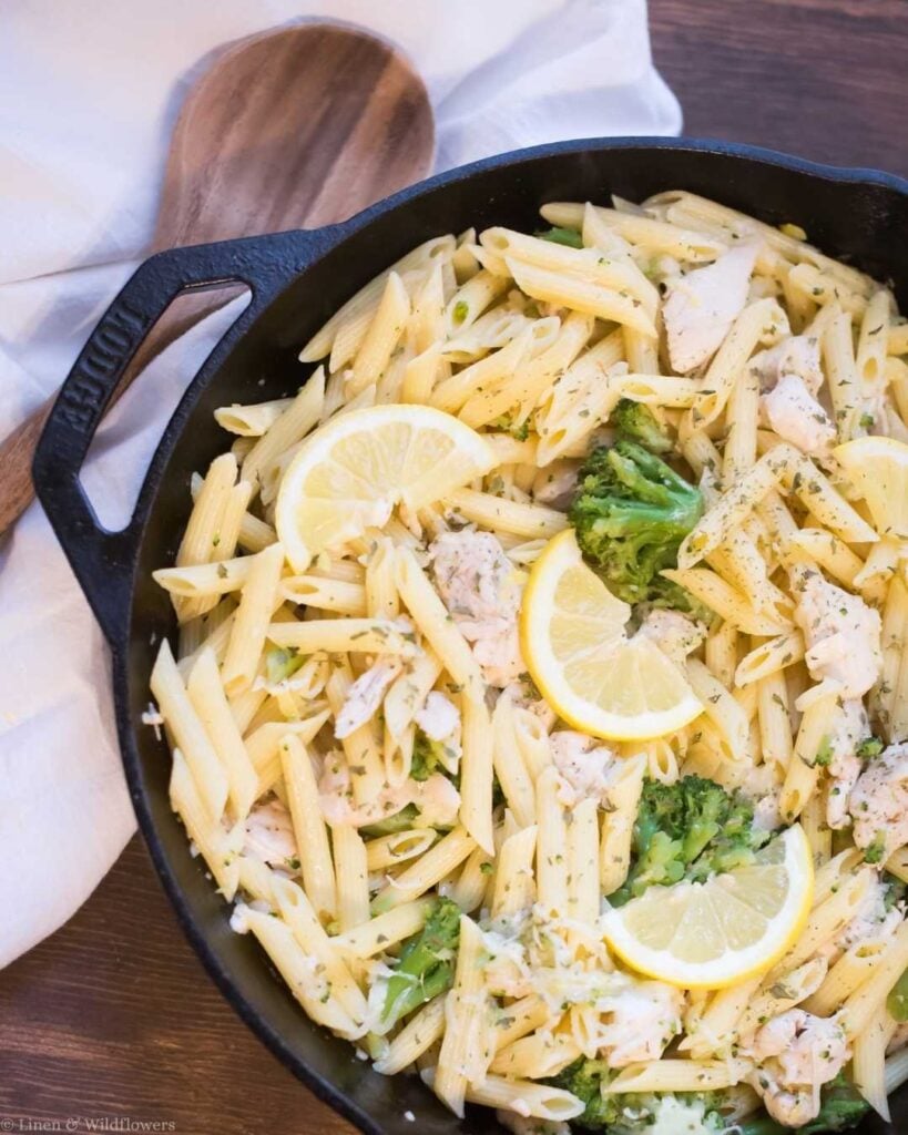Cast iron skillet filled with penne pasta, broccoli, chicken, and lemon slices on a wood table — budget-friendly frugal homemaker dinner idea to save money at home