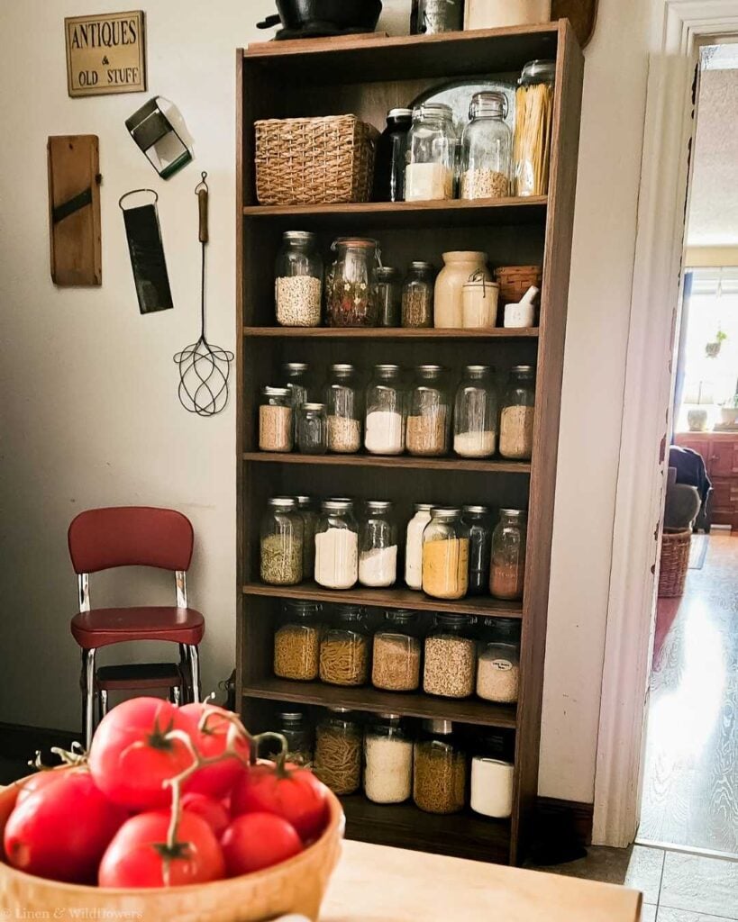 Organized homemaker pantry shelf stocked with glass mason jars of grains, pasta, and dry goods with a bowl of fresh tomatoes — how to save money at home