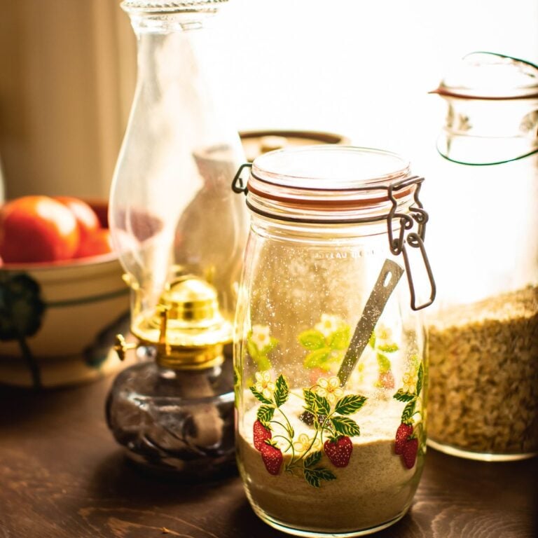 Vintage strawberry print glass jar filled with flour beside grain jars and an oil lamp in warm kitchen light — frugal homemaker pantry staples to save money at home