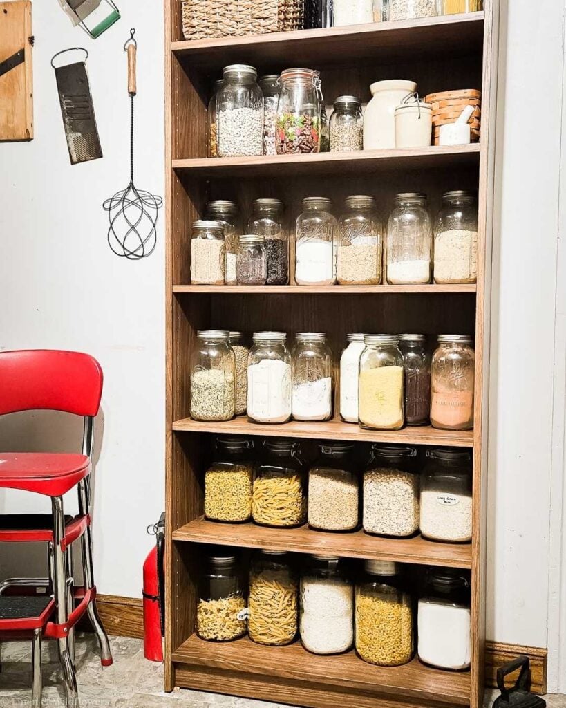 Tall wooden pantry bookshelf fully stocked with glass mason jars and clip-top jars storing pasta, grains, flour, and dry goods on every shelf
