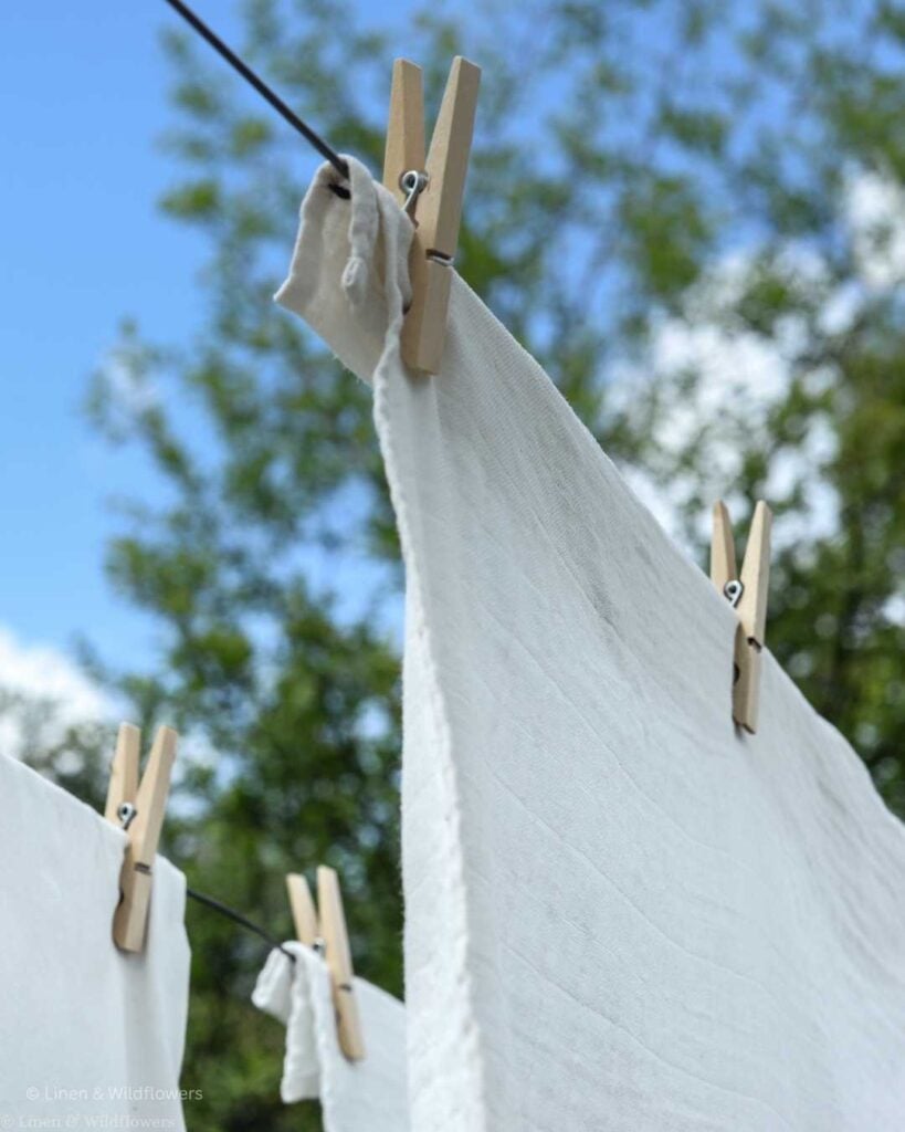 White linens hanging on an outdoor clothesline with wooden clothespins against a blue sky and green trees — frugal homemaker tip to save money on energy bills