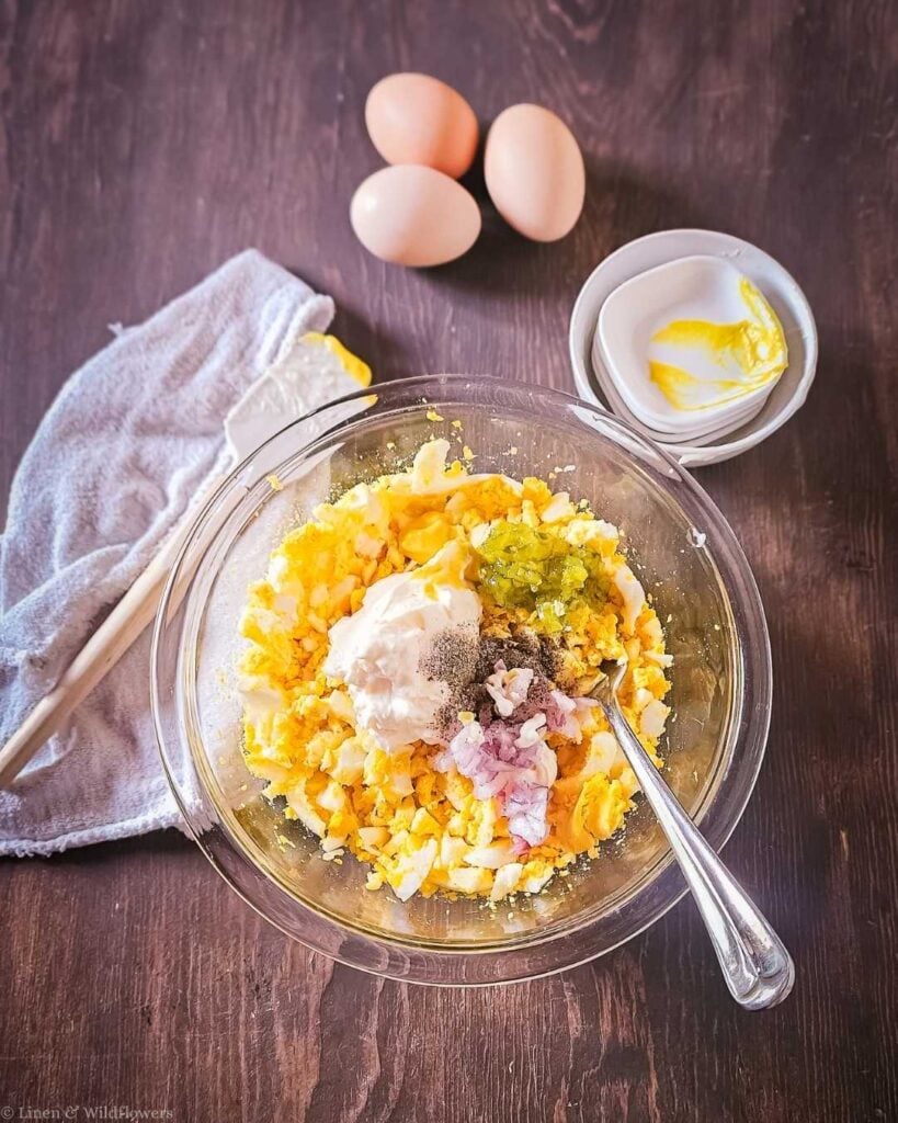 Glass bowl of chopped hard-boiled eggs topped with mayonnaise, sweet relish, diced red onion, and black pepper before mixing for classic egg salad.