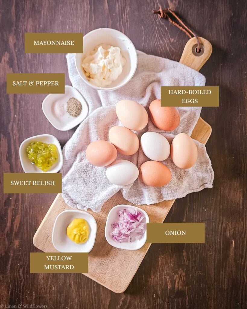 Flat lay of old-fashioned egg salad ingredients on a wooden cutting board, including hard-boiled eggs, mayonnaise, yellow mustard, sweet relish, diced red onion, salt, and pepper.
