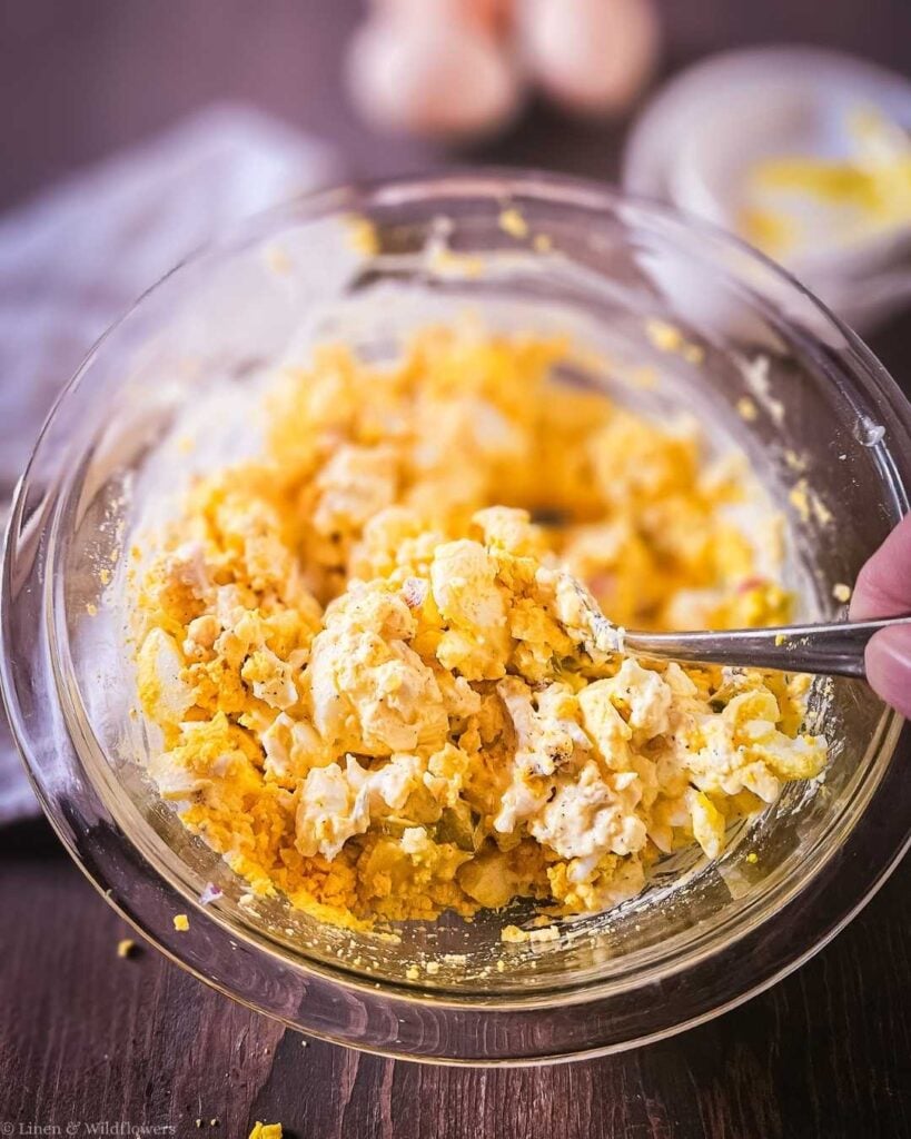 Hand mixing old-fashioned egg salad with a fork in a glass bowl, combining chopped eggs with mayonnaise, mustard, and relish.