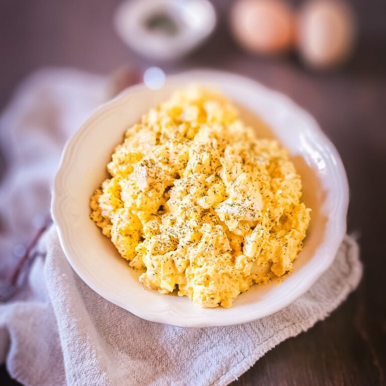 Creamy old-fashioned egg salad with dried dill served in a white scalloped bowl on a linen napkin, with hard-boiled eggs and seasoning blurred in the background.