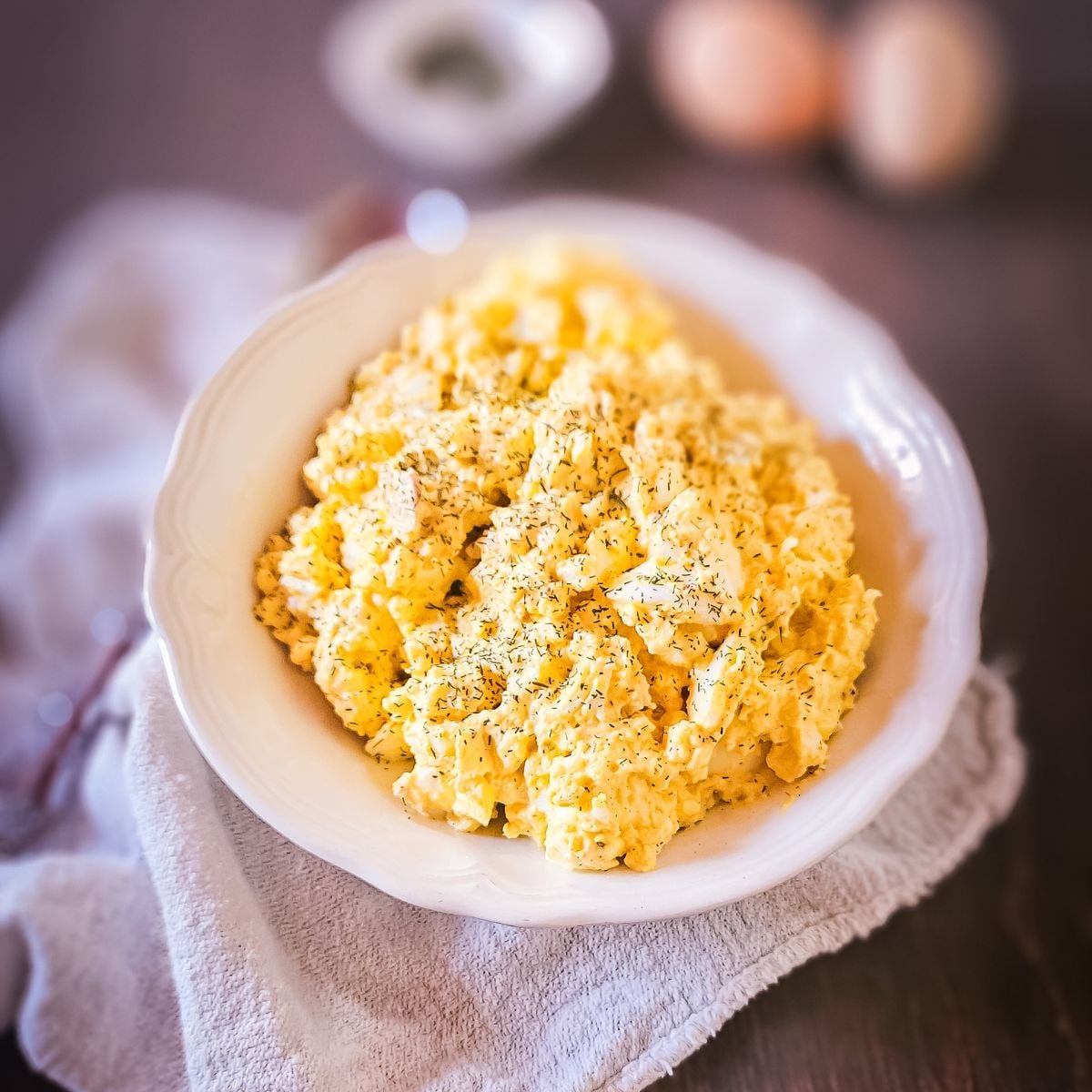 Creamy old-fashioned egg salad with dried dill served in a white scalloped bowl on a linen napkin, with hard-boiled eggs and seasoning blurred in the background.