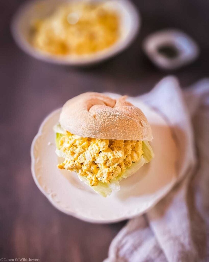 Old-fashioned egg salad sandwich served on a crusty dinner roll with crisp lettuce on a white plate, with a bowl of egg salad blurred in the background.