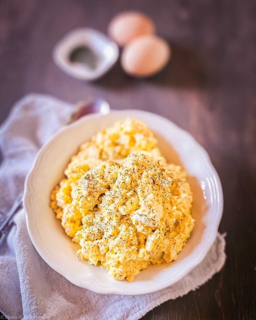 Creamy old-fashioned egg salad served in a white scalloped bowl, garnished with dried dill, on a rustic dark wood surface.