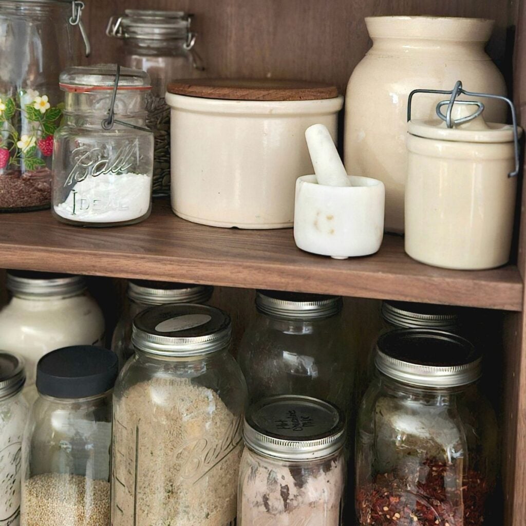 Pantry shelf detail featuring vintage stoneware crocks, a marble mortar and pestle, Ball Ideal glass jar with sugar, and rows of mason jars filled with grains, hot chocolate mix, and dried herbs below