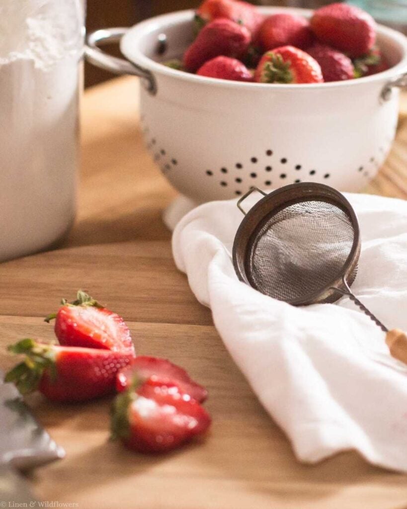 Fresh strawberries in a white colander with a fine mesh strainer and linen cloth on a wood cutting board — frugal homemaker cooking from scratch