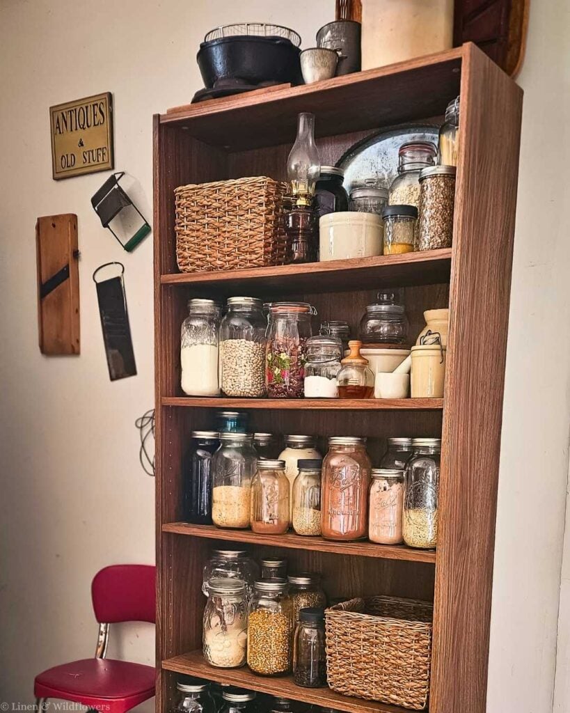 Full view of a vintage farmhouse pantry bookshelf filled with glass mason jars of dry goods, wicker baskets, stoneware, and cast iron cookware on top, with antique wall decor beside it