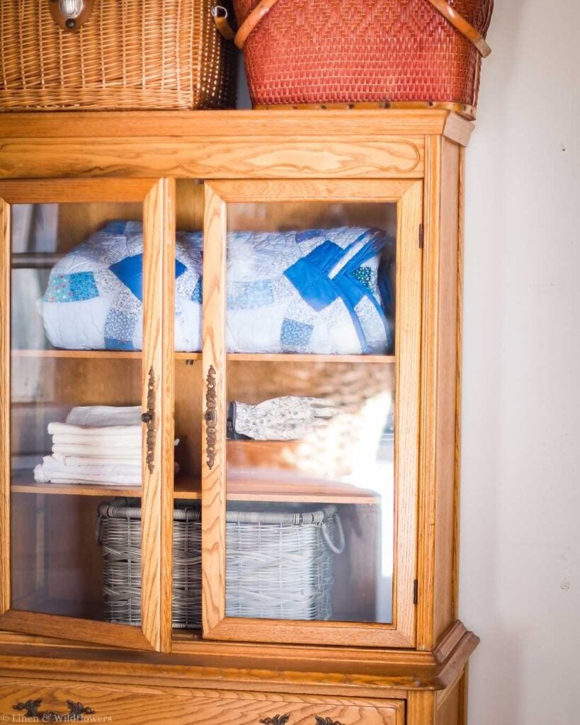 Vintage storage ideas using an antique oak glass-front cabinet to display folded quilts, linens, and wicker baskets.