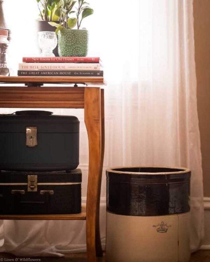 Vintage storage idea using stacked train cases under a wooden side table next to an antique stoneware crock.
