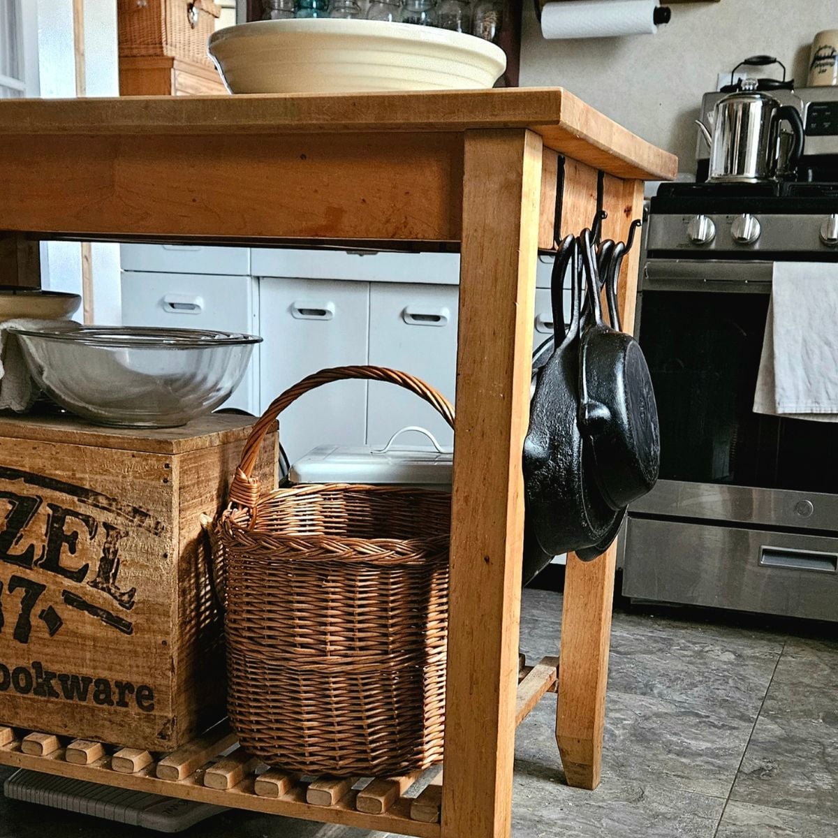 Vintage storage idea using an antique wood crate, wicker basket, and hanging cast iron pans for charming kitchen island organization.