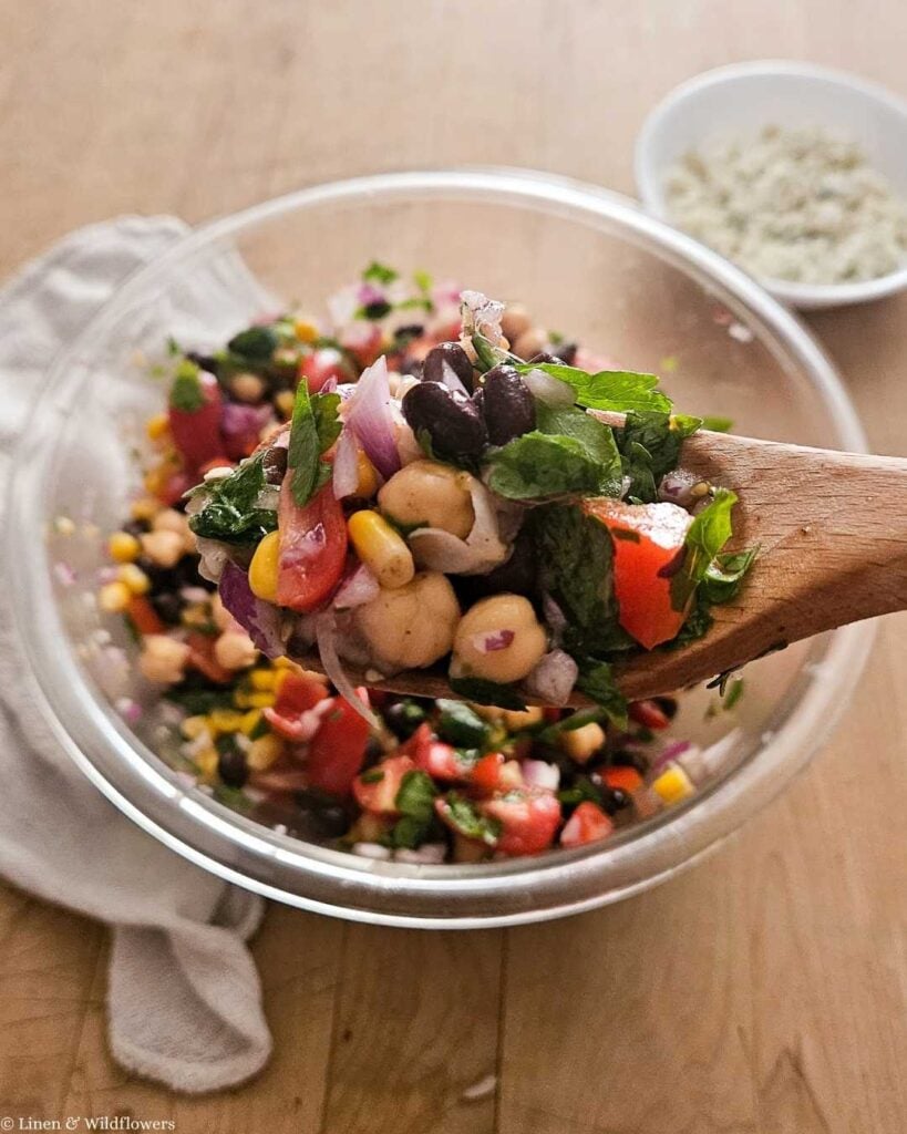 Wooden spoon lifting a scoop of chickpea salad with lemon, beans, tomato, and herbs.