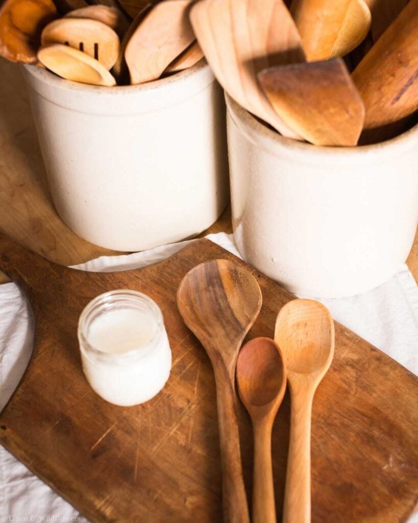 Wooden spoons beside a jar of oil on a cutting board, demonstrating how to oil wooden spoons to prevent damage.