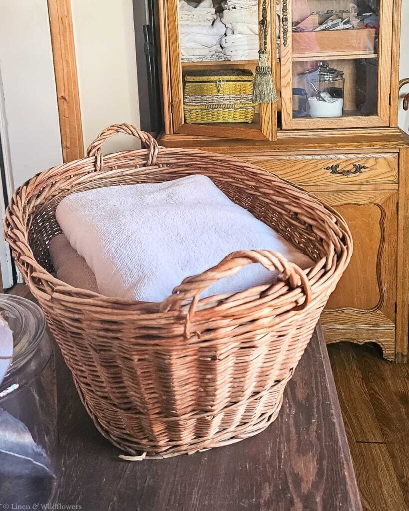 Large vintage wicker laundry basket with woven handles holding folded white towels on a farmhouse table.