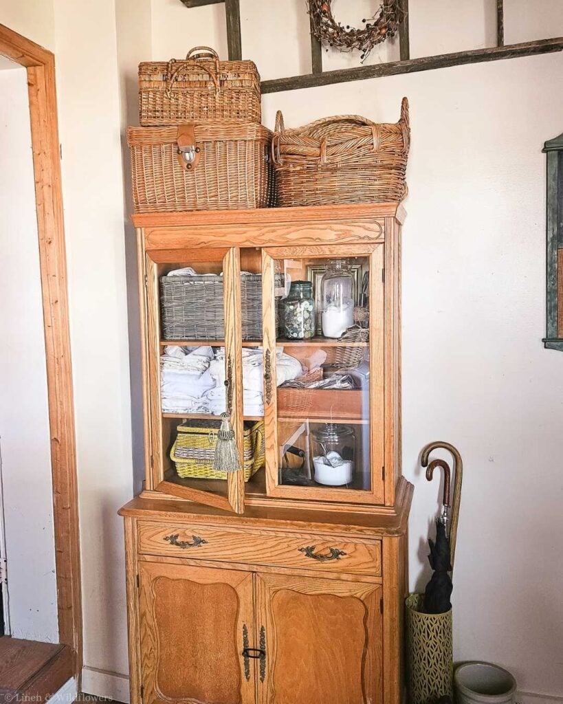Collection of vintage wicker baskets, including a lidded picnic basket, displayed on top of an oak china cabinet.