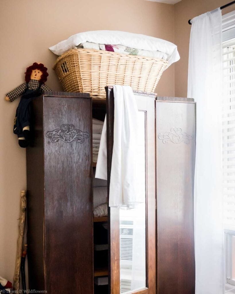 : Vintage wicker basket filled with folded linens sitting atop an antique wooden wardrobe.