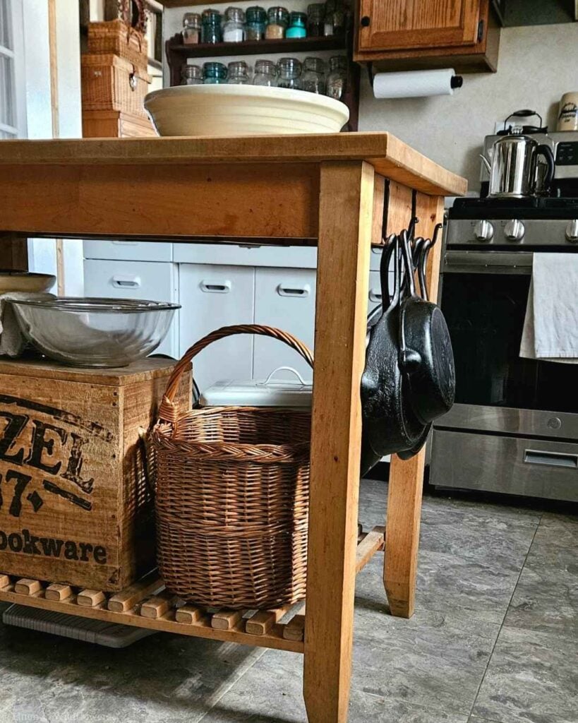 Butcher block kitchen island with a wicker basket, vintage crate, and cast iron skillets hanging from hooks.