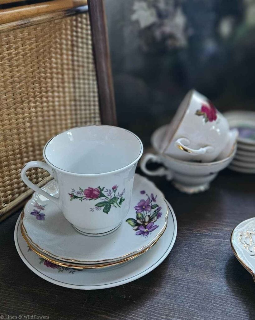 Thrifted vintage floral teacups and saucers stacked on a wicker chair, budget-friendly kitchen charm.