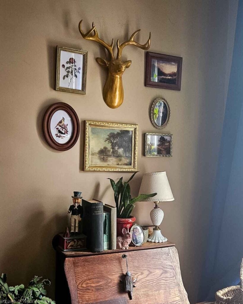 Styled vintage gallery wall vignette with gold deer head, framed botanical and landscape art, and thrifted decor atop an antique desk.