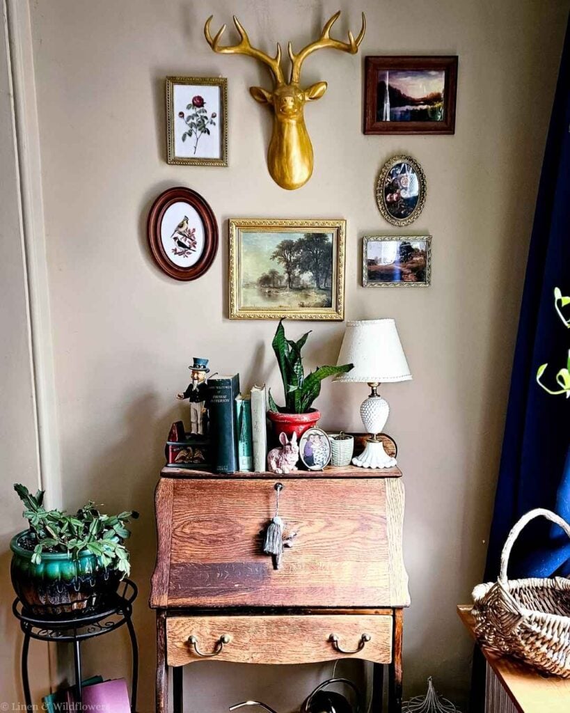 Finished vintage gallery wall with gold deer head, botanical prints, and landscape paintings arranged above an antique oak secretary desk.