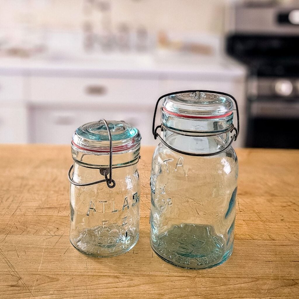 Two vintage Atlas glass jars with wire bail closures and red rubber replacement seals on a wood counter.