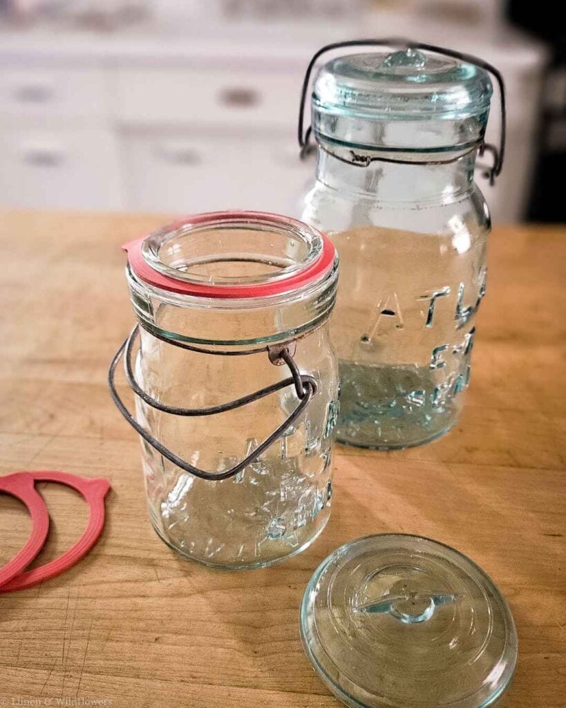 Vintage glass canning jars with wire bail closures and red rubber sealing rings on a wood counter.