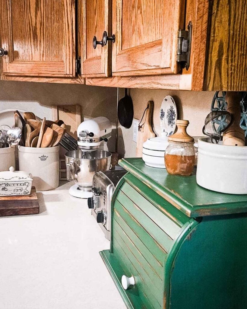 Distressed green bread box, stoneware crocks, and butter dish styled on a farmhouse kitchen counter.