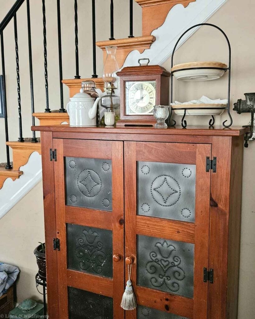 Antique pie safe styled with a vintage clock, oil lamp, and ironstone teapot on top.