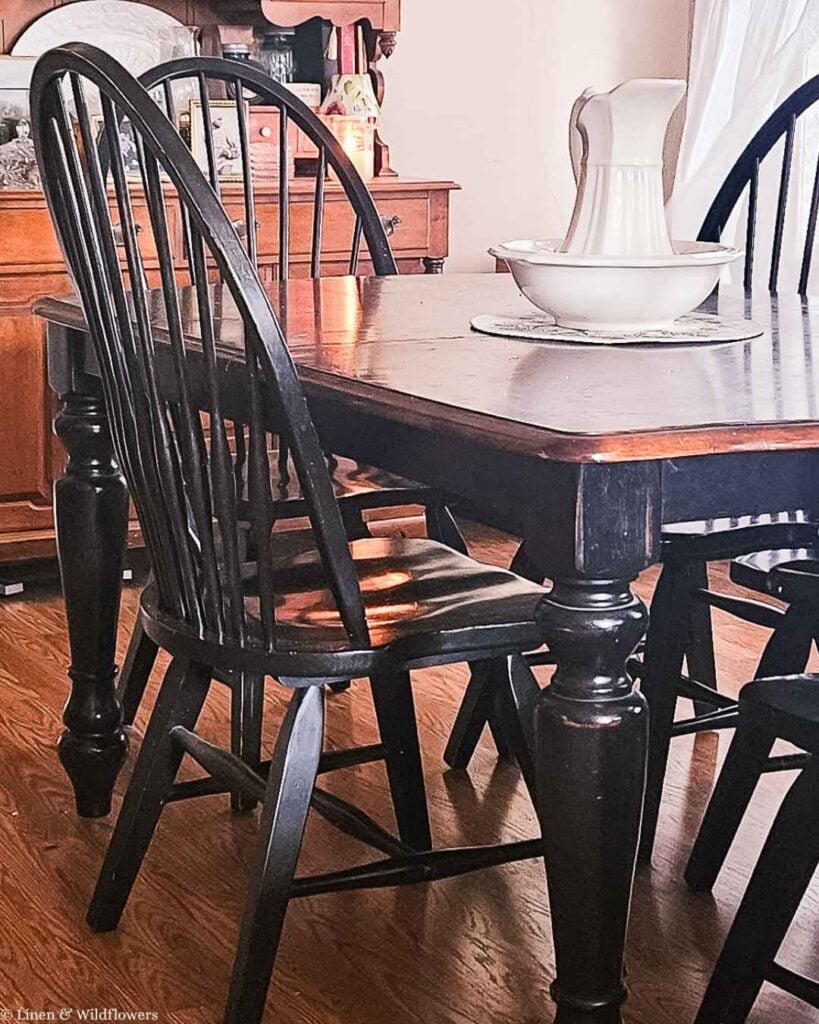 : Close up of black Windsor chair spindle back next to farmhouse dining table with ironstone pitcher.
