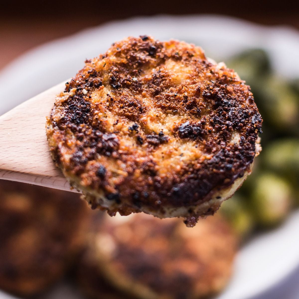 Crispy fried eggplant parmesan round on a wooden spoon, served with brussels sprouts.