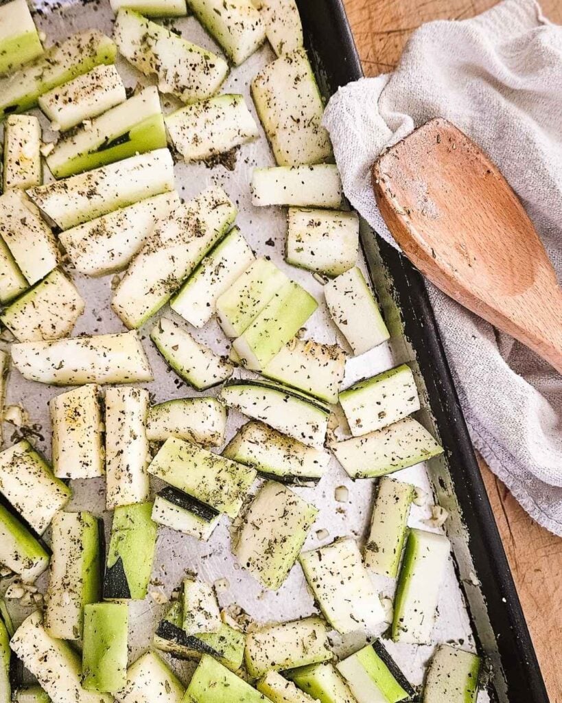 Close-up of herb-seasoned zucchini pieces on a sheet pan before roasting, with a wooden spoon.