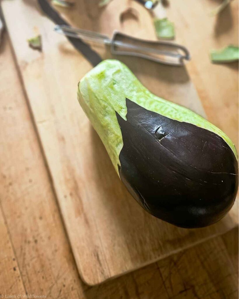 Peeling the skin off a fresh eggplant with a vegetable peeler on a cutting board.