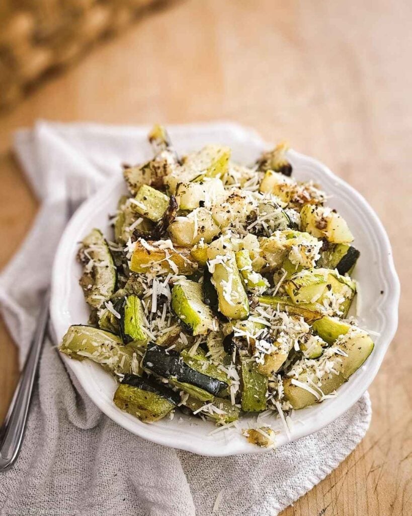 A bowl of roasted garlic Parmesan zucchini garnished with fresh Parmesan and cracked black pepper.