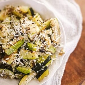 Close-up of roasted garlic Parmesan zucchini in a white bowl, topped with melted Parmesan cheese and cracked black pepper.
