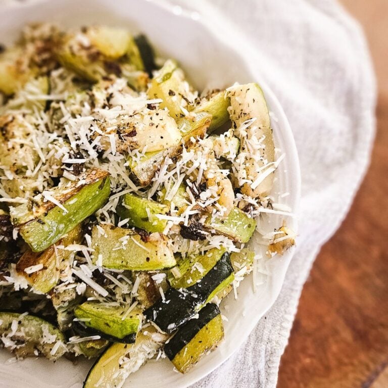 Close-up of roasted garlic Parmesan zucchini in a white bowl, topped with melted Parmesan cheese and cracked black pepper.