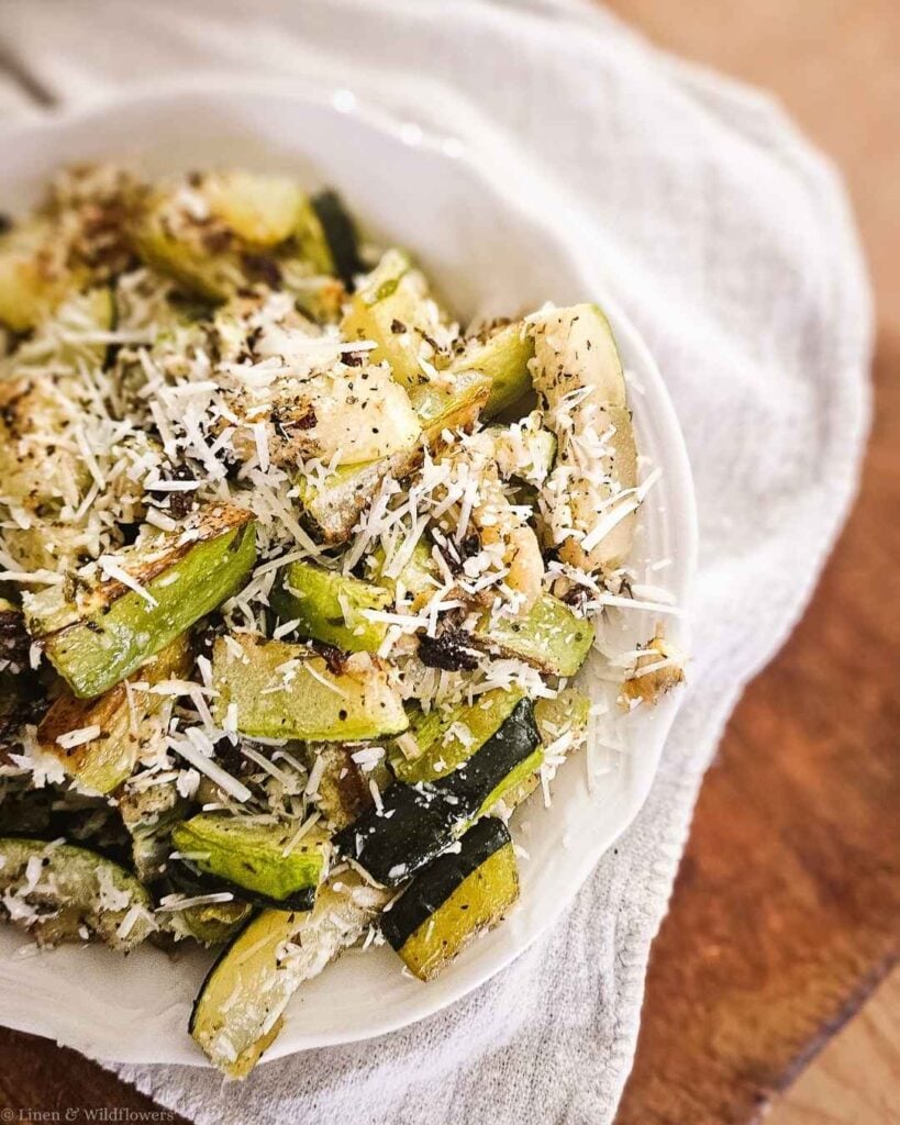 Close-up of crispy roasted zucchini tossed with garlic and Parmesan cheese in a white serving bowl.