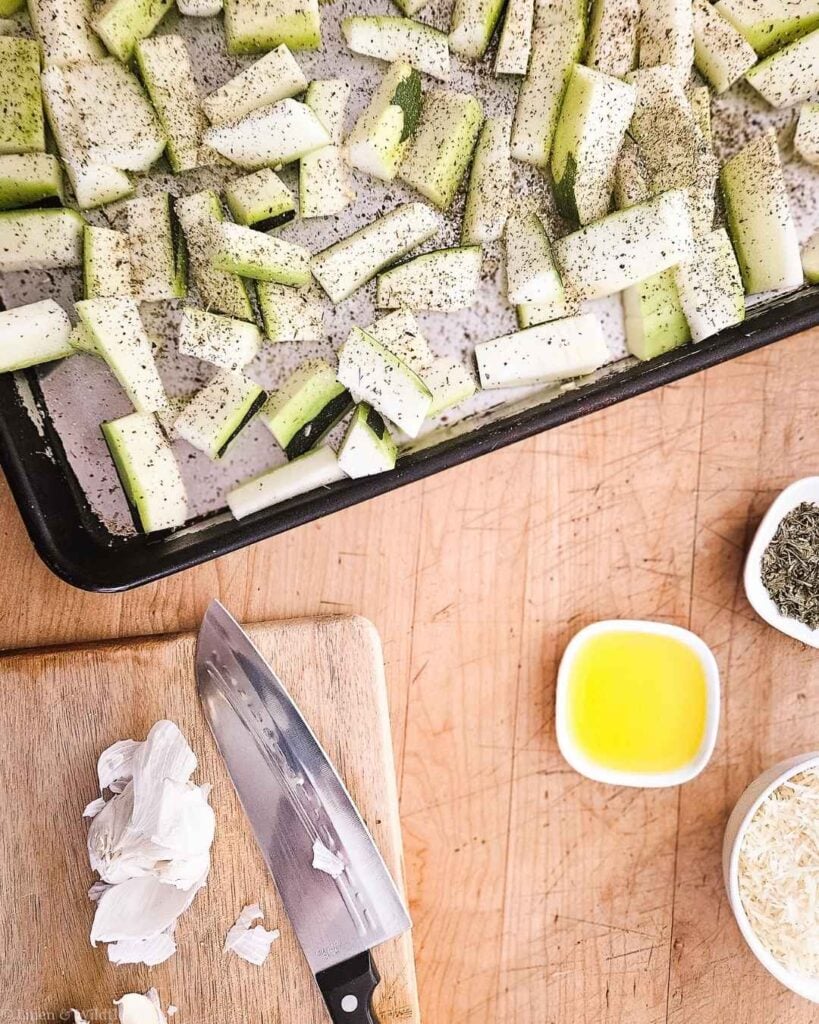 Zucchini strips seasoned with herbs and pepper on a baking sheet, with garlic, olive oil, and Parmesan nearby.