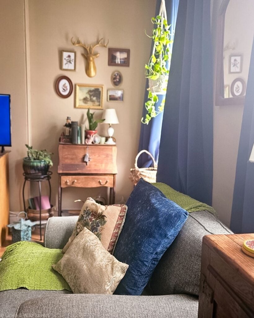 Cozy living room mixing a small wooden secretary desk, framed gallery wall, and vintage lamp with blue curtains.