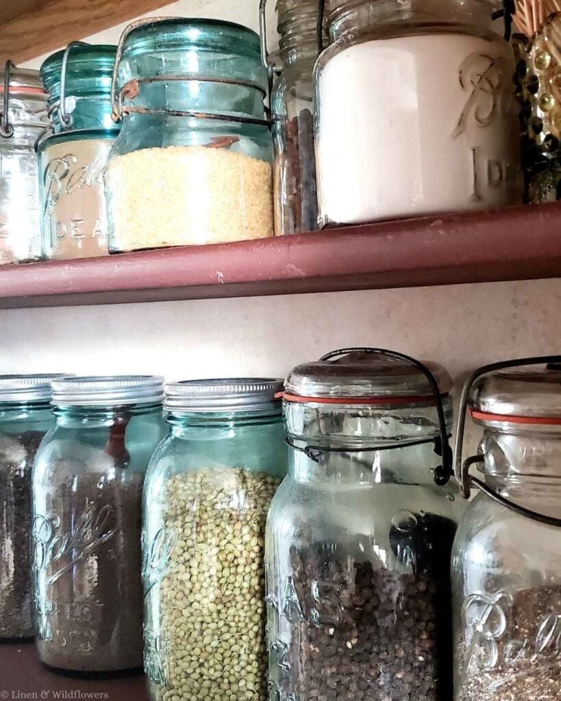 Antique blue Ball canning jars holding couscous, coriander seeds, and peppercorns on a painted pantry shelf.