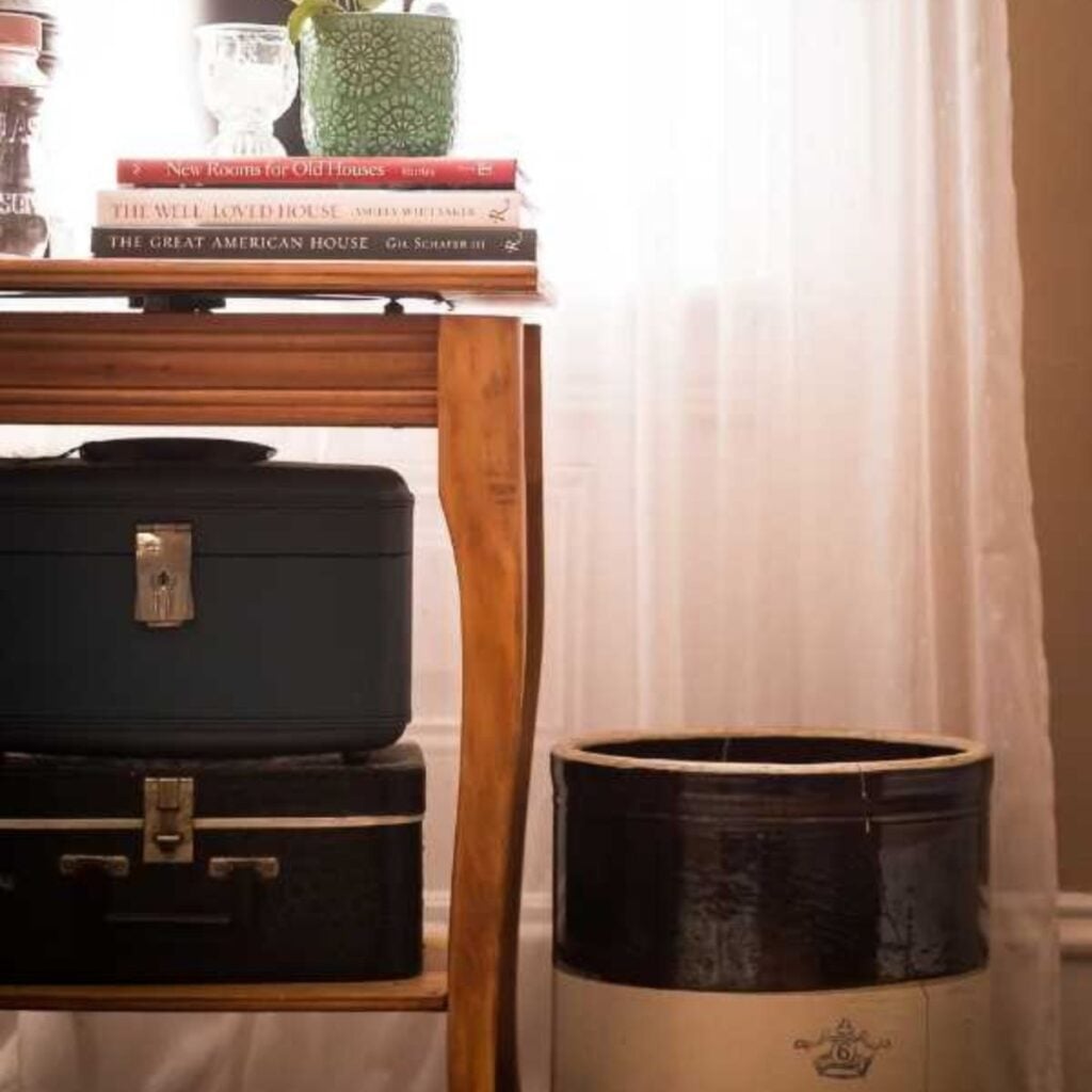 Stoneware crock beside a wooden table holding stacked vintage train cases and a stack of home decor books with a green planter