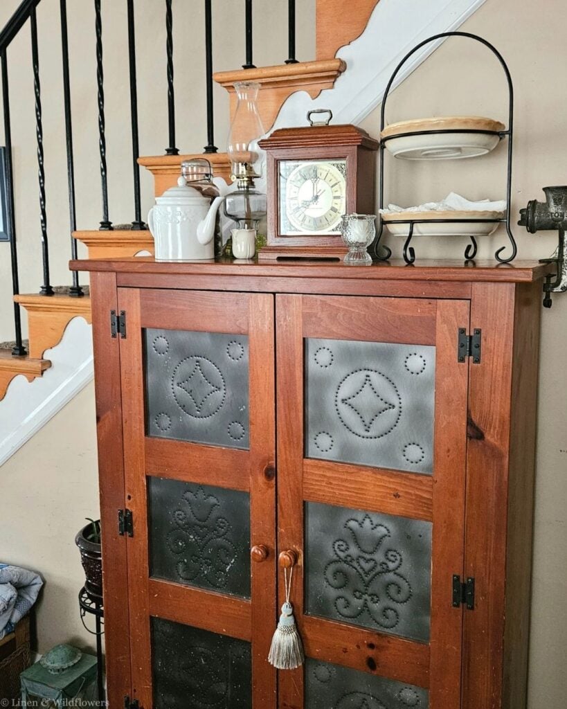 Antique pine pie safe with punched tin panels styled with a mantel clock, oil lamp, and ironstone teapot.