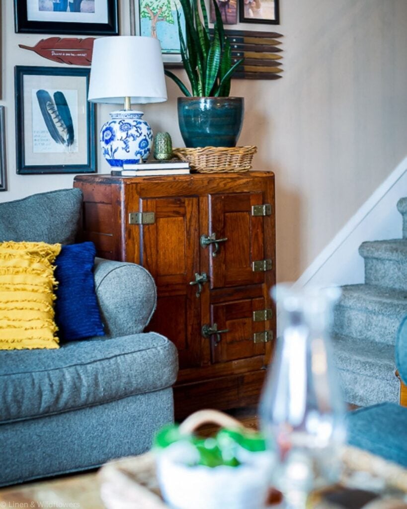 Wooden ice box cabinet with brass hinges styled with a blue and white lamp, plant, and basket in a cozy living room.
